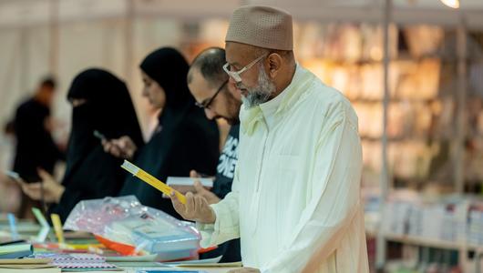 A man at a book fair reads the back of a book he is holding.